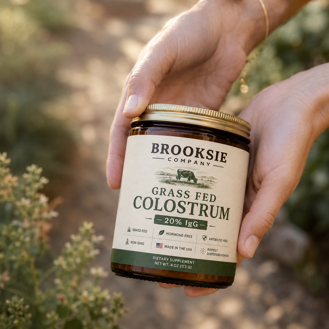 A person's hands holding a jar of Brooksie Company Grass Fed Colostrum against a blurred outdoor background.