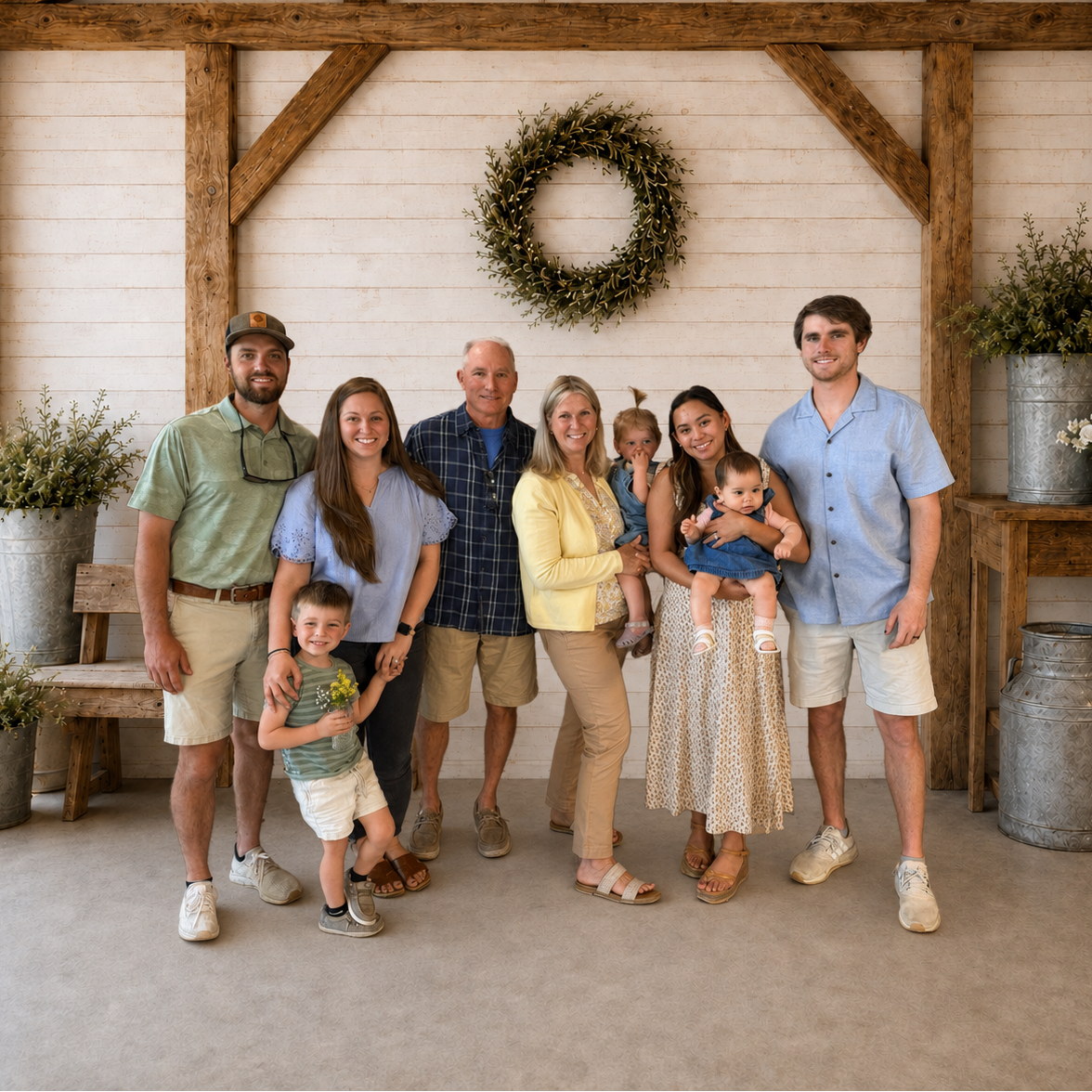 A family of seven posing together in a rustic barn interior with white wood paneling, exposed wooden beams, a green wreath on the wall, and potted plants. The group includes a man, a woman, a toddler, and two children wearing casual summer clothing.